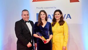 A man in a suit and two women in a blue dress and yellow dress stood in a line at an awards ceremony.  The women in the blue dress is holding an award