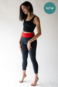 A lady with back curly hair standing in a photography studio, smiling and looking otwards the floor.  She is wearing a pair of Davy J Sustainable Waterwear black swim leggings with the waist band folded down to show the red lining.  She is also wearing a black cropped swim top.