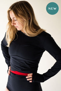 A lady with blonde hair, stood with her hands on her hips and looking down towards the floor.  She is wearing a black Davy J Sustainable Waterwear long sleeve black top and high waisted black and red bikini briefs