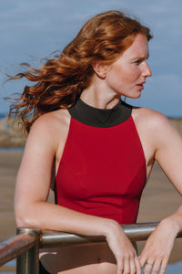 A lady with red hair leaning with her arms on a hand rail, looking out to the distance.  She is wearing a red high necked Davy J Sustainable waterwear swim top with black neck detail
