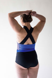 The back view of a lady with short brown hair stood in a photography studio with her arms outstretched above her head.  She is wearing a Davy J Sustainable Waterwear black and blue crossback swim top and black and blue high waisted bikini briefs 