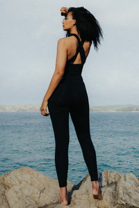 The rear view of a lady with black afro hair, standing on a rock and looking out to sea.  She is wearing a Davy J sustainable swimwear black crossback swim top and black swim leggings