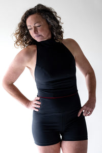 A lady with short brown curly hair, stands with her hands on her hips and looks down at the floor.  She is wearing a black Davy J sustainable waterwear high neck swim top and black high waisted swim shorts