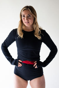 A lady with blond hair, stood in strong stance with her hands on her hips, looking straight towards the camera.  She is wearing a Davy J Sustainable Waterwear long sleeve black swim top and black high waist bikini briefs with a red turned down waistband 