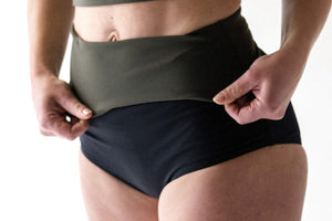 A close up detail of a lady wearing a pair of black high waisted swim briefs and folding down the waist to show the contrasting olive coloured lining