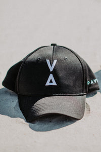 A Davy J Sustainable Waterwear black cotton baseball cap laid on sand.  The cap has a white embroidered Davy J logo on the front and a white Davy J text logo embroidered on the side