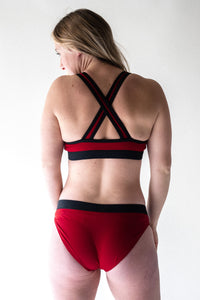 Back view of a blonde lady standing up looking over her shoulder wearing a Davy J Sustainable Waterwear red bikini top with a crossback and black elastic detail and red bikini briefs with a black waistband 
