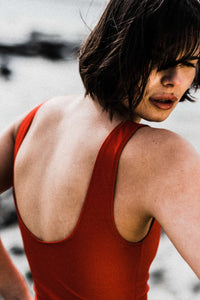 A lady with black bobbed hair looking over her right shoulder.  She is wearing a red Davy J Sustainable Waterwear cropped swim top with a scoop back