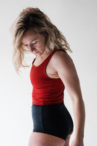 A side view of a blonde lady with her arm in the air, wearing a Davy J Sustainable Waterwear red cropped swim top with scoop back and shoulder straps.  She also wears a pair of black high waist bikini briefs