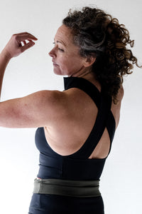 Woman in studio turning back towards camera with arm raised wearing black Davy J Sustainable Waterwear high neck halter swim top with wide cross back straps and black high waist bikini briefs rolled down to show olive green lining