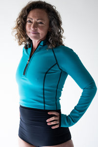 A lady with brown curly hair smiling has her hands on her hips.  She wears a Davy J Sustainable Swimwear ocean green long sleeve swim top with th zip partially undone and a pair of high waist black bikini briefs