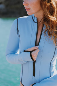 A close up view of a lady with auburn hair standing in front of the sea, wearing a Davy J Sustainable Waterwear powder blue long sleeve swimsuit, pulling up the zip detail with her right hand