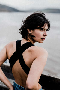 Woman with brunette short hair facing away to the sea, wearing jeans over her black Davy J Sustainable Waterwear cutout swimsuit with wide cross back straps 