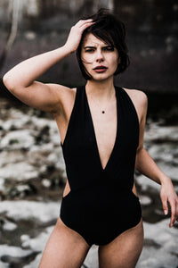 Woman stood on beach frowning with one hand on head, wearing black Davy J Sustainable Waterwear cutout swimsuit with plunging neckline 