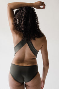 Back view of woman with long curly brown hair and one arm raised in studio wearing an olive green Davy J Sustainable Waterwear cutout swimsuit with wide cross back straps