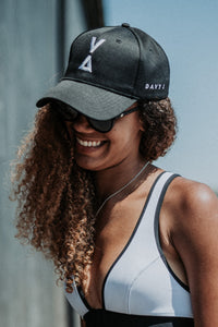 A girl smiling with long brown curly hair is wearing a Davy J Sustainable Waterwear white bikini top with black edging.  She is also wearing black sunglasses and a black Davy J cotton baseball cap with white Davy J logo embroidered on the front and a white Davy J text logo embroidered on the side