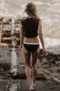 A slim blonde lady walks down a slipway towards the sea.  She wears a Davy J Sustainable Waterwear black short sleeve swim top with black low rise bikini briefs with a white waistband