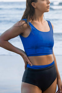 Woman stood on shoreline looking away with one hand on waist, wearing Davy J Sustainable Waterwear blue crop swim top and dark grey high waist bikini briefs rolled down to show blue lining. 