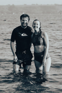 A woman and man stood in the sea smiling and looking straight at the camera