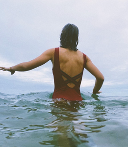 A back view of a lady with dark hair walking through water with her arm outstretched, wearing a Davy J Sustainable Waterwear red swimsuit with crossback
