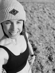 Black and white image of a woman in a black Davy J Sustainable Waterwear swimsuit and wearing a beanie hat on a beach smiling