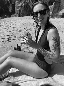 A woman sat on a towel on a beach in a Davy J Sustainable Swimwear cutout swimsuit wearing big sunglasses with lots of arm tattoos