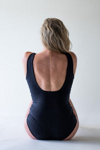 A rear view of blonde lady sitting on the floor in a photography studio, with her hair tucked over her shoulder.  She is wearing a black Davy J sustainable waterwear low backed swimsuit
