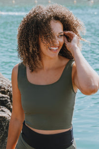 A lady with brown afro hair, stood infront of the sea smiling.  She is wearing an olive Davy J Sustainable Waterwear swim top
