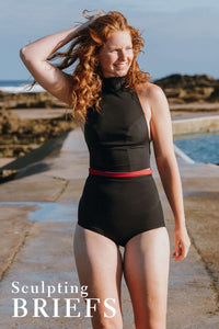 A lady with red hair walking down the side of an outdoor swimming pool, wearing a black swim top and black high waisted bikini briefs.  There is a text overlay on the image which says 'Sculpting briefs'.