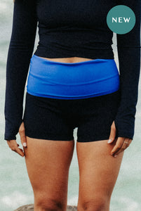 A lady standing in front of the sea wearing a pair of Davy J Sustainable waterear black high waisted swim shorts with a blue turned down waistband.  She is also wearing a black long sleeved swim top.