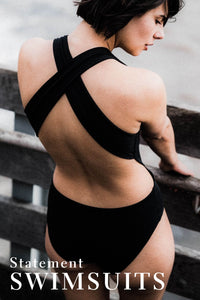 A lady with short black hair and wearing a black crossback swimsuit, leaning against a wooden fence.  There is a text overlay on the image that says 'Statement Swimsuits'