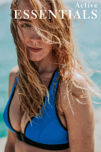 A lady with blonde hair blowing across her face looks at the camera with the sea behind her.  She is wearing a blue bikini top.  There is a text overlay on the image that says 'Active Essentials'.