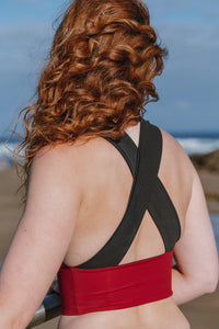 The back of a lady with curly red hair, looking out to the sea.  She is wearing a black and red Davy J sustainable waterwear crossback swim top
