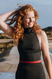 A lady with red hair smiling and looking out to sea, with her arm outstretched, playing with her hair.  She is wearing a black Davy J Sustainable waterwear swim top with black high waisted bikini briefs