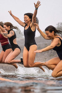 A group of ladies smiling and wearing Davy J Sustainable Waterwear swimsuits, jumping into the sea with arms outstretched 