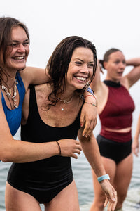 A lady stood in the sea wearing a black Davy J Sustainable Waterwear black swimsuit.  Another lady wearing a blue swim top has her arm wrapped around her shoulder 
