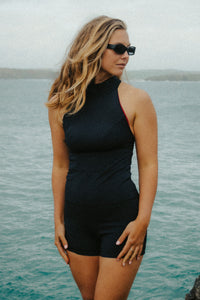 A tanned lady with blonde hair wearing black sunglasses, standing in front of the sea and looking over her right shoulder.  She is wearing a Davy J sustainable waterwear black high neck swim top and black high waisted swim shorts  