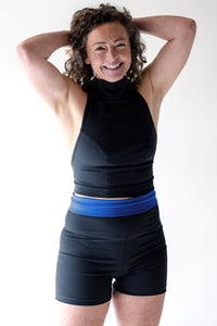 A lady with short brown curly hair has both her hands reaching up towards her head.  She is smiling and looking into the camera and wearing a black Davy J sustainable waterwear high neck swim top and black swim shorts with a blue waistband