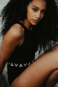A close up image of a lady with long black afro hair, crouched down looking directly into the camera.  She is wearing a black Davy J sustainable swimsuit with a black and white logo belt. 