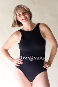 A lady with short hair standing with her hands on her hips, looking directly into the camera and smiling.  She is wearing a black Davy J sustainable waterwear swimsuit with a black and white logo belt. 