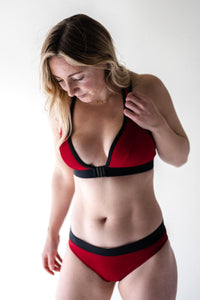 A blonde lady looking down to the floor wearing a Davy J Sustainable Swimwear red bikini top with black front opening clasp and red low rise bikini briefs with black waistband