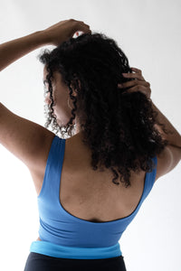 A back view of a lady with her hands running through her black curly hair, wearing a Davy J Sustainable Waterwear blue cropped swim top with scoop back and shoulder straps