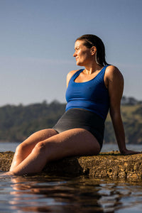 A lady sat on a stone wall with her legs in the sea, looking out at the water wearing a Davy J Sustainable Waterwear blue cropped swim top with squared neckline and shoulder straps and black high waist briefs