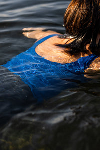 Close up view of the back of a woman swimming in clear water, wearing a Davy J Sustainable Waterwear blue cropped swim top with squared back and grey high waisted bikini briefs