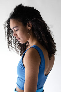 A side facing image of a lady with long black curly hair and big hooped earring, wearing a Davy J Sustainable Waterwear blue cropped swim top with scooped back and shoulder straps 