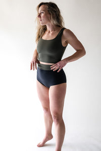 A lady with blonde hair stood on tip toes, wearing a Davy J Sustainable Waterwear olive cropped swim top with squared neckline and shoulder straps.  Worn with a pair of black high waisted bikini briefs with the waist rolled down showing a band of olive lining
