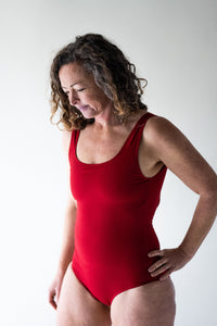 Woman with brown curly hair in studio looking downwards with hand on hip wearing Davy J Sustainable Waterwear red classic crossback swimsuit 
