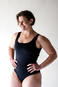 Smiling woman with short brunette bob with hands on hip in studio, wearing a Davy J Sustainable Waterwear black classic cross back swimsuit
