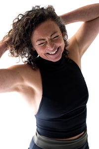 Laughing woman in studio with hands in her brown curly hair wearing black Davy J Sustainable Waterwear high neck halter swim top and black high waist bikini briefs rolled down to show olive green lining