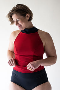 Smiling woman with short brown hair gazing downwards  in studio wearing Davy J Sustainable Waterwear red halter swim top with black collar overlapping black high waist bikini briefs rolled down to show red lining. 